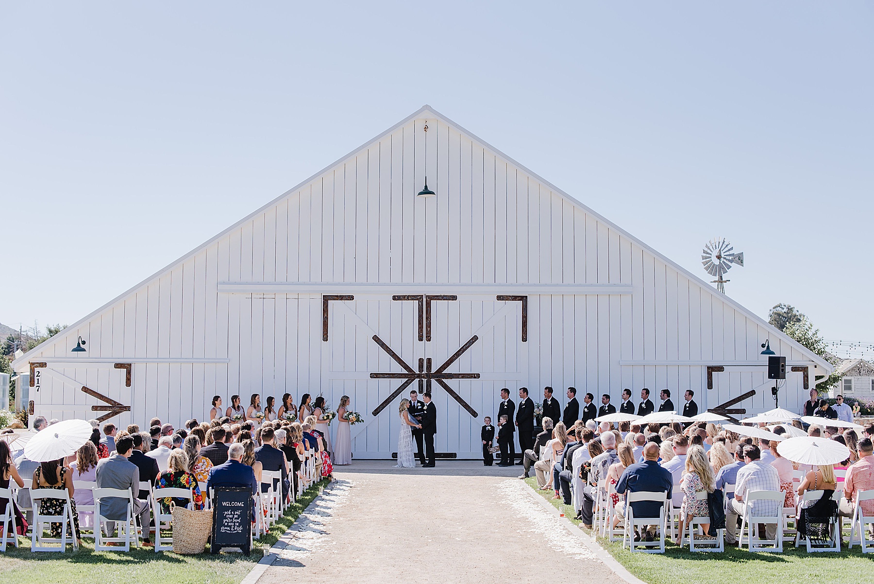 Wedding ceremony in front of a white, rustic barn. Wedding guests are seated. The wedding party is standing on both sides of the bride and groom.