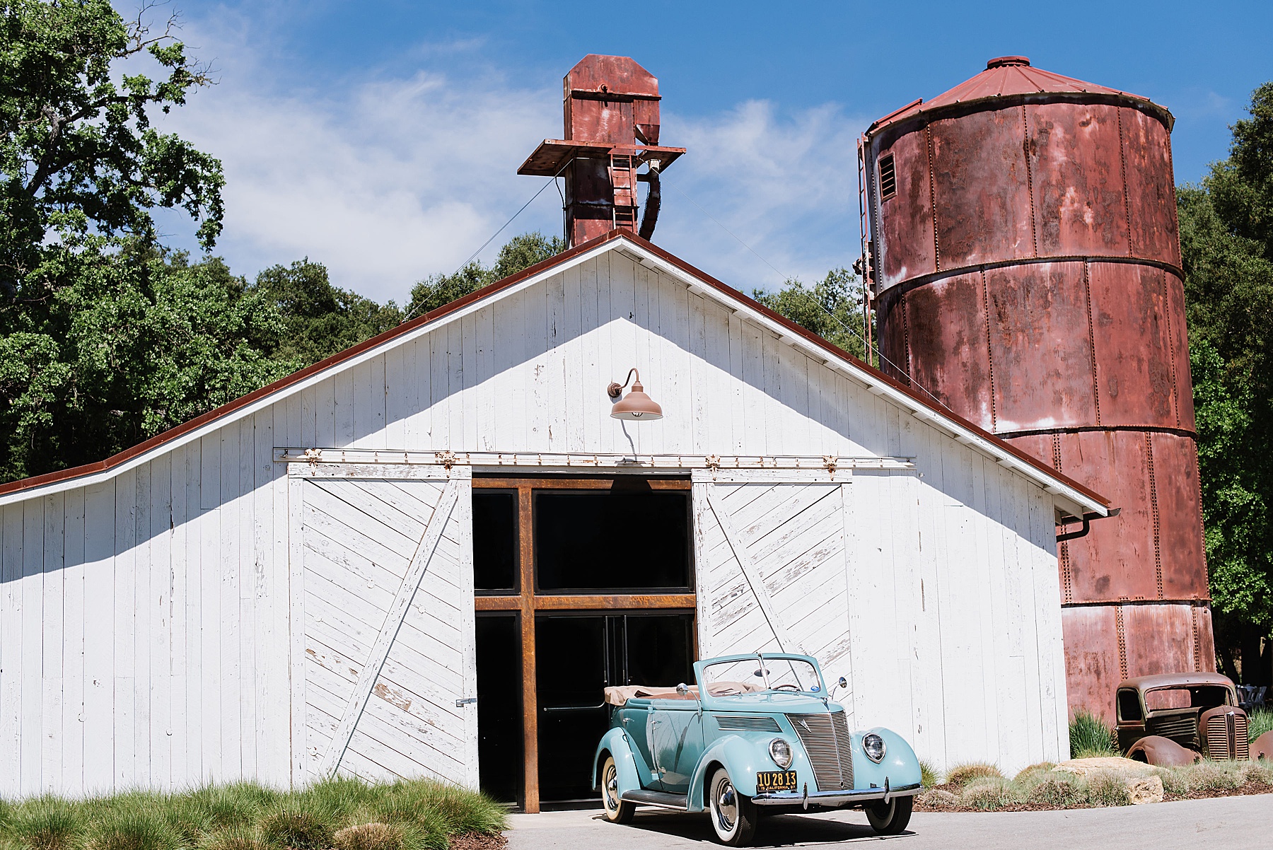 White, rustic barn that is next to a red silo. A old, vintage, blue car sits out front.
