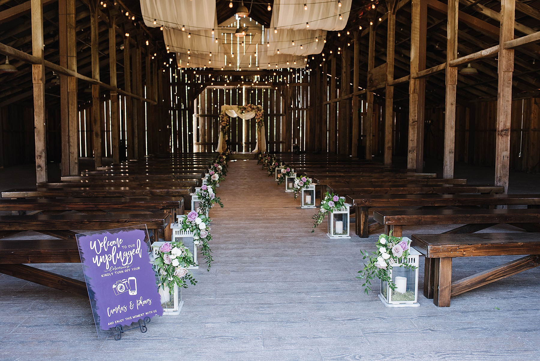 Rustic wedding venue. String lights are lining the ceiling. White lanterns with pink floral accents line the aisle.