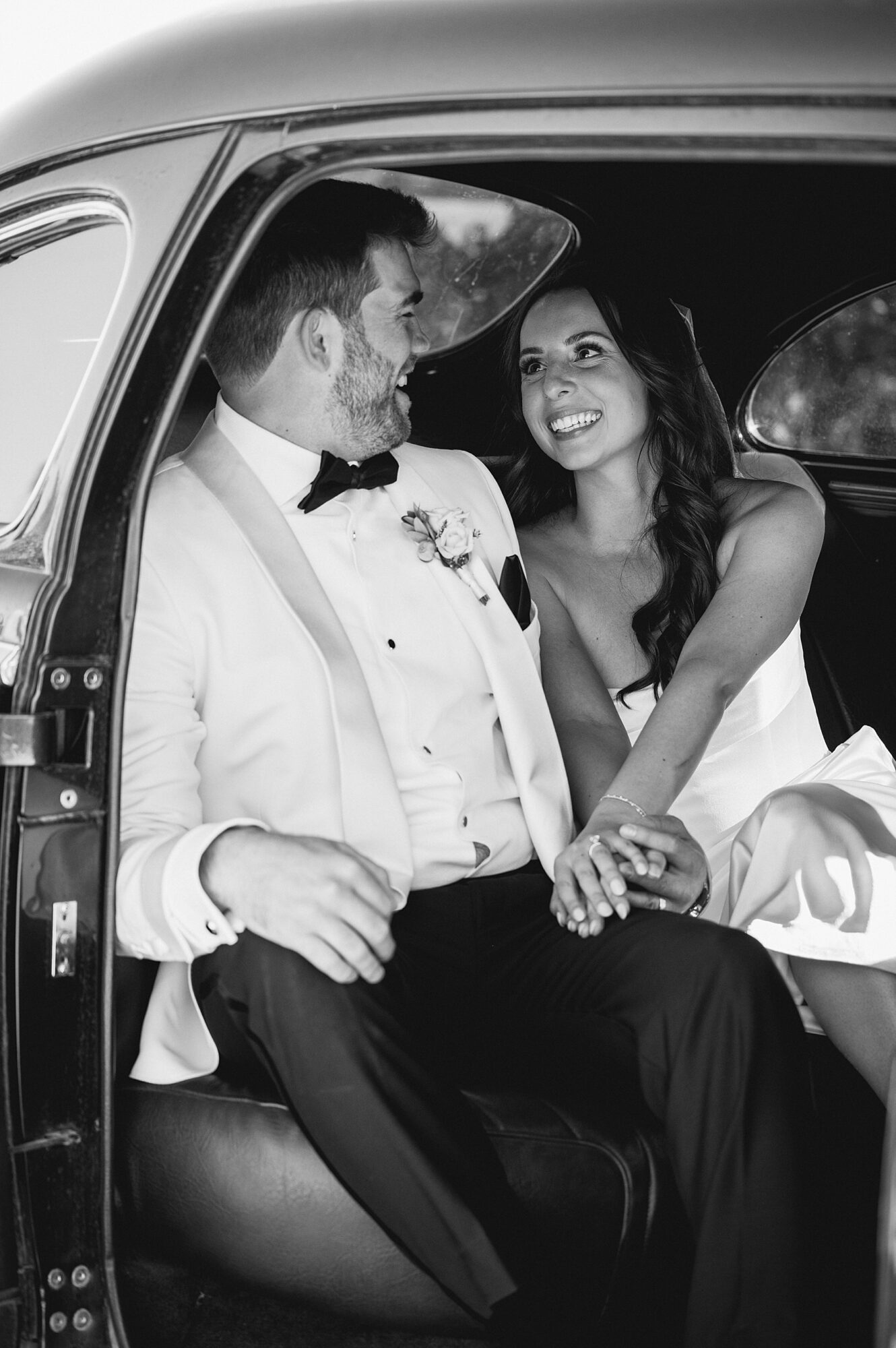 Black and white photo of a joyful bride and groom sitting inside a vintage car. The groom is smiling at the bride, who is beaming as they hold hands, capturing an intimate and happy moment after their wedding ceremony.