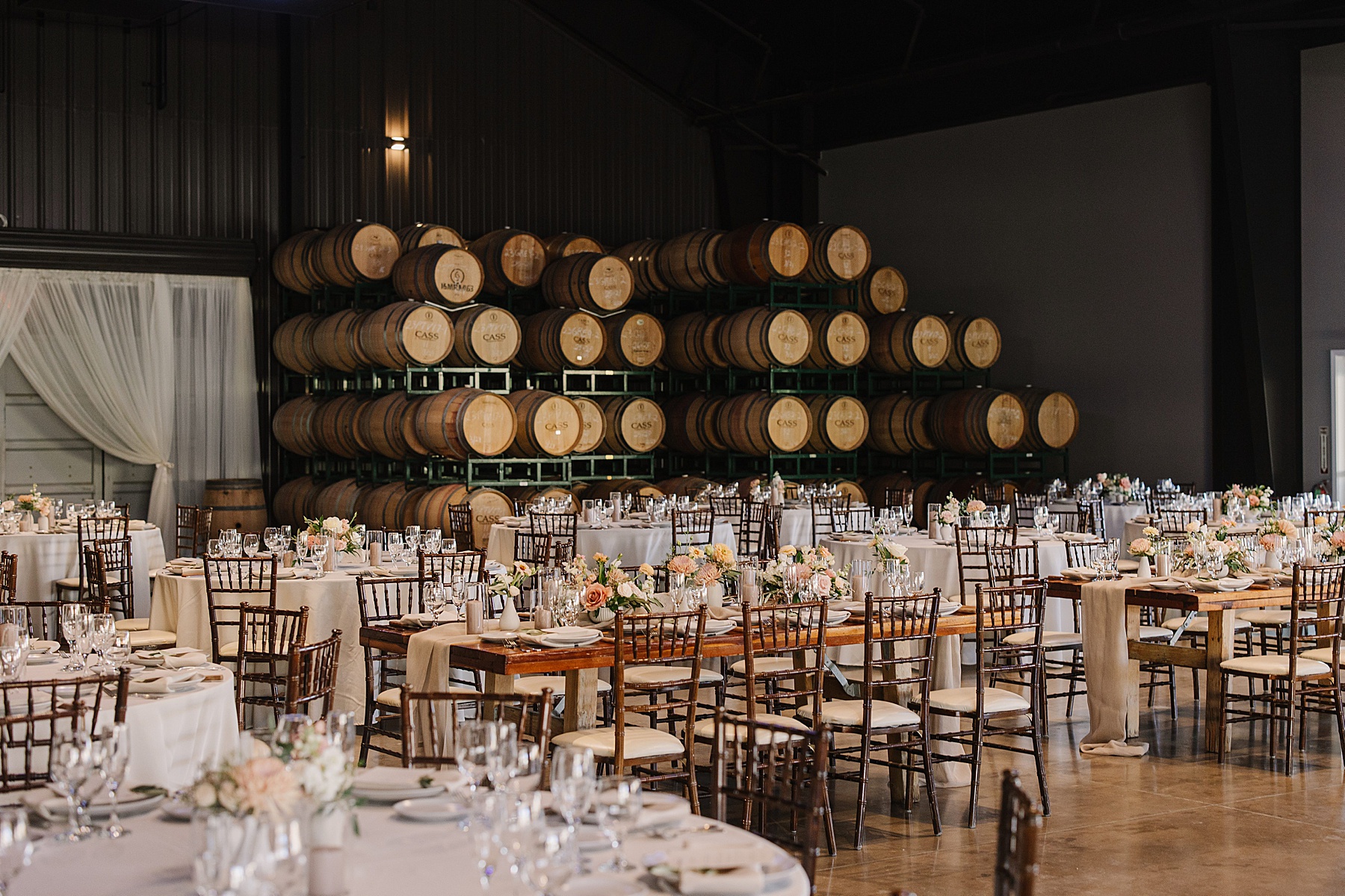 Elegant wedding reception setup inside the barrel room at Cass Winery in Paso Robles, featuring round and rectangular tables with floral centerpieces, set against a backdrop of stacked wine barrels. The tables are adorned with glassware, plates, and soft, neutral table runners, creating a rustic yet refined atmosphere