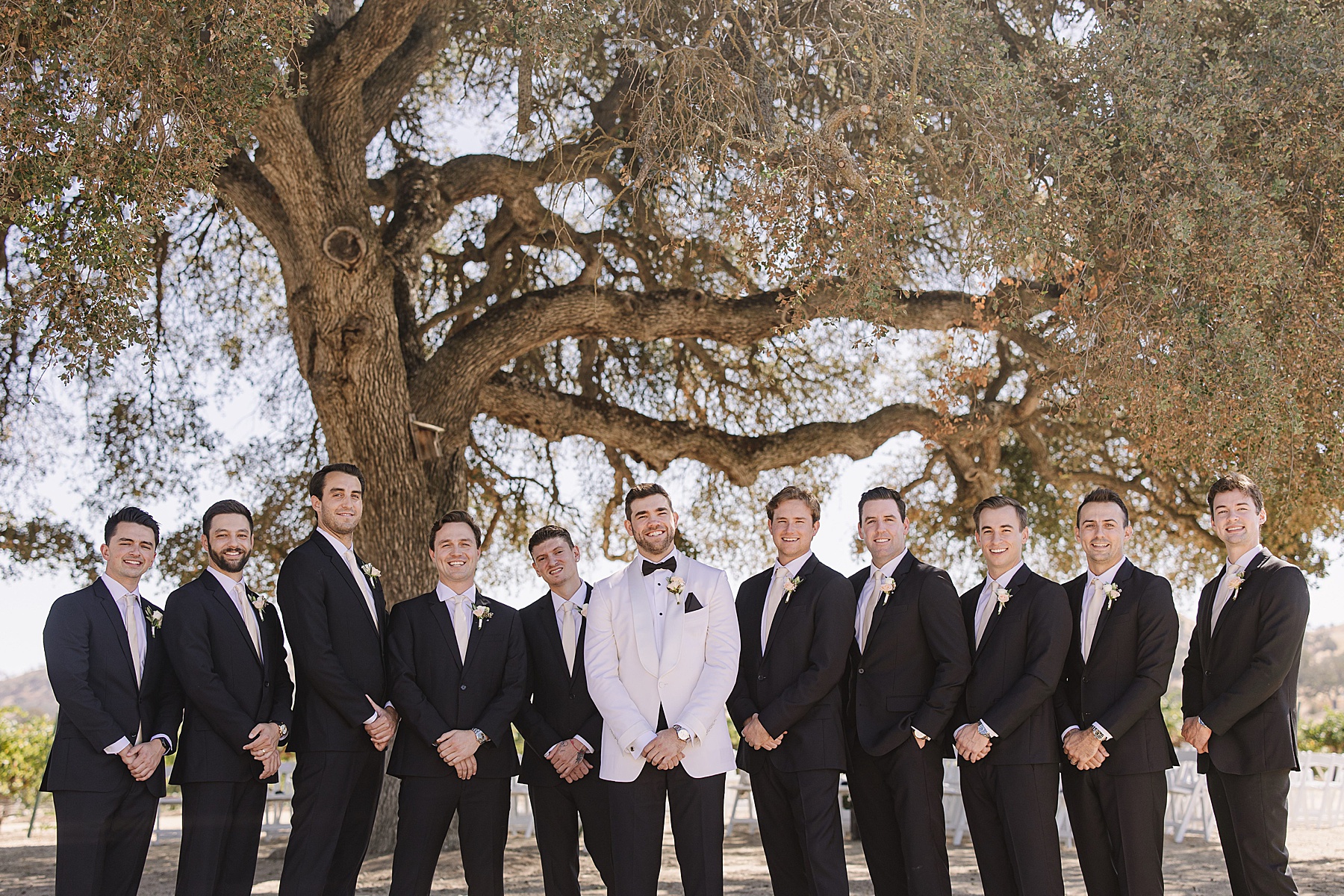 Groom standing with his groomsmen under a large tree at Cass Winery in Paso Robles. The groom is dressed in a white tuxedo jacket, while the groomsmen wear classic black suits with matching boutonnieres. The group smiles together, with the vineyard and rustic landscape visible in the background.