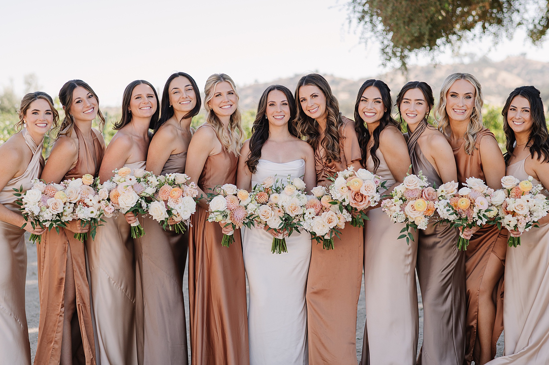 Bride standing with her bridesmaids, all smiling and holding bouquets of soft peach and cream flowers. The bridesmaids are wearing elegant satin dresses in shades of champagne, bronze, and taupe, complementing the natural backdrop of Cass Winery in Paso Robles.