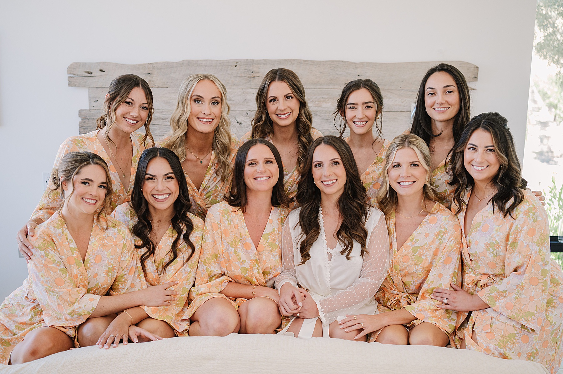 Bride surrounded by her bridal party, all wearing matching floral robes, as they sit together on a bed. The bride is in the center wearing a white robe, while the bridesmaids wear peach-colored robes with a daisy pattern. They are all smiling in a cozy, indoor setting.