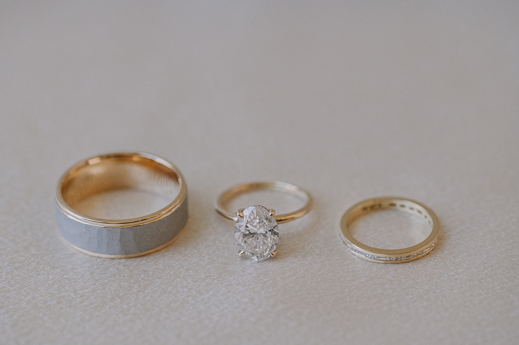 Close-up of three wedding rings arranged side by side on a light-colored surface: a wide gold band, an engagement ring with a large oval diamond, and a slim gold band with small inset diamonds.