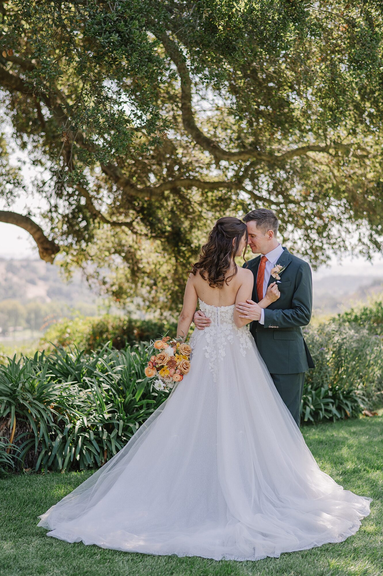 Romantic bride and groom portrait at Foxen Canyon Ranch, with the couple embracing under the shade of oak trees, surrounded by lush greenery and the bride holding a vibrant bouquet of orange and yellow blooms.