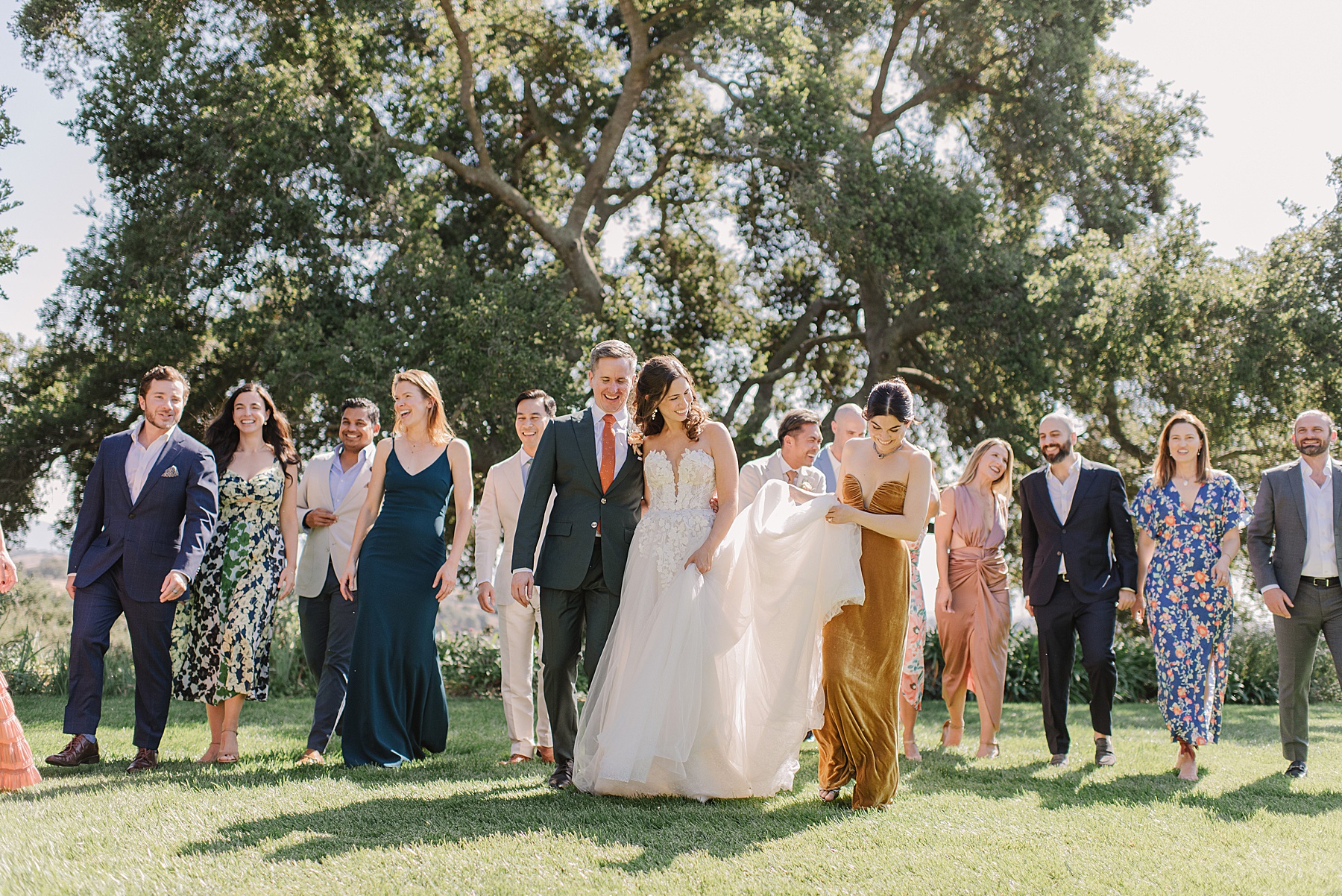 Vibrant wedding party strolling together at Foxen Canyon Ranch, with the bride and groom at the center, surrounded by friends in colorful attire, under the shade of majestic oak trees.