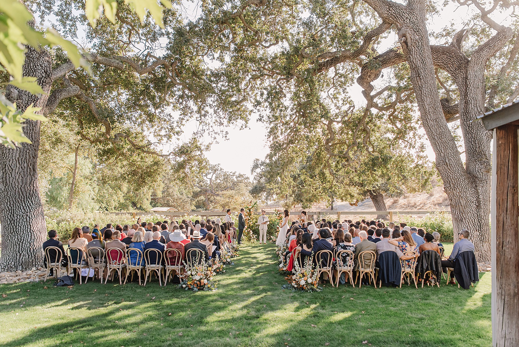 Enchanting outdoor ceremony at Foxen Canyon Ranch, with the couple exchanging vows under the canopy of ancient oak trees, surrounded by family, friends, and lush greenery.