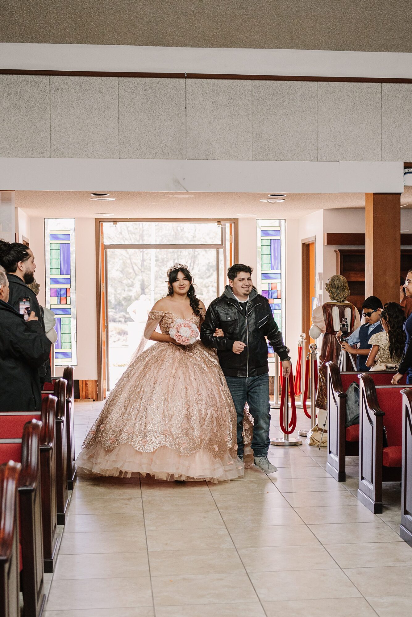 Mary Luz walks into the church with her escort during her Tooth + Nail Winery Quinceañera, wearing a stunning blush gown adorned with intricate details.