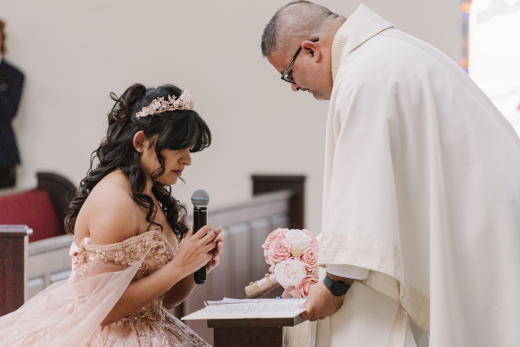 Mary Luz kneels during a heartfelt moment of her Tooth + Nail Winery Quinceañera ceremony, holding a microphone and her elegant bouquet."