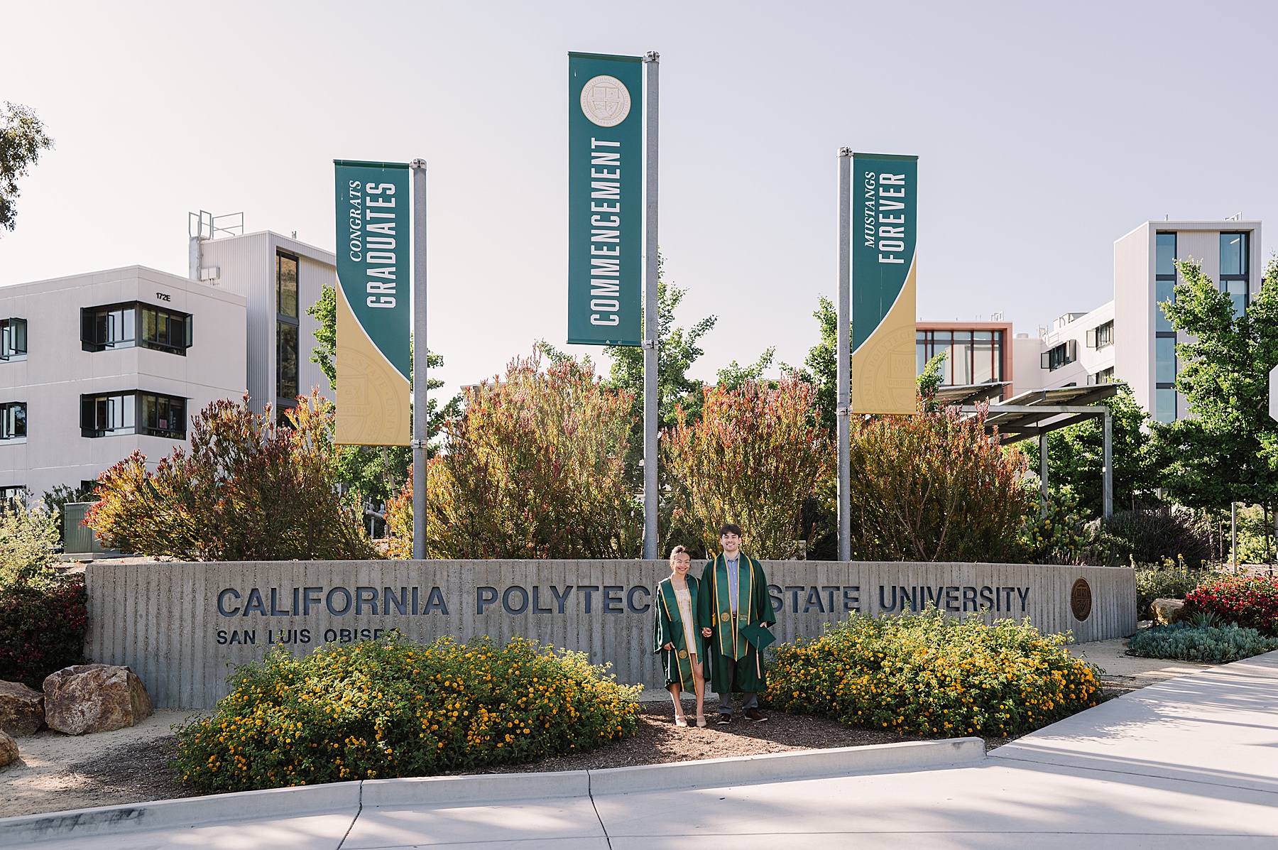 Two graduates in green and gold gowns posing in front of the California Polytechnic State University sign in San Luis Obispo, celebrating their Cal Poly senior photos session with banners reading 'Congrats Graduates' and 'Mustangs Forever' in the background.