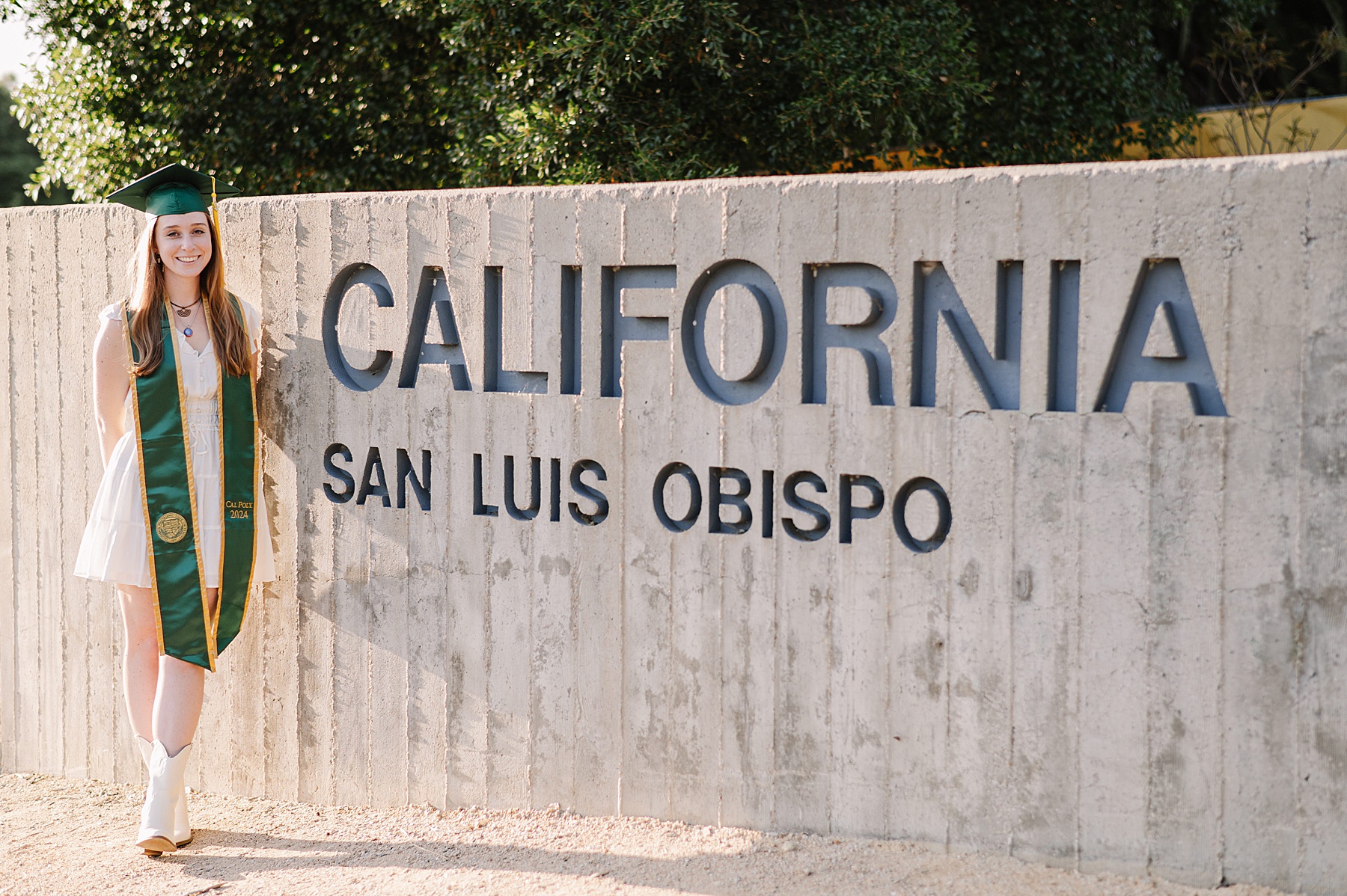 A graduate in a white dress and cowboy boots poses beside the California Polytechnic State University sign in San Luis Obispo during her Cal Poly senior photos session, wearing a green and gold stole with the class of 2024 emblem.