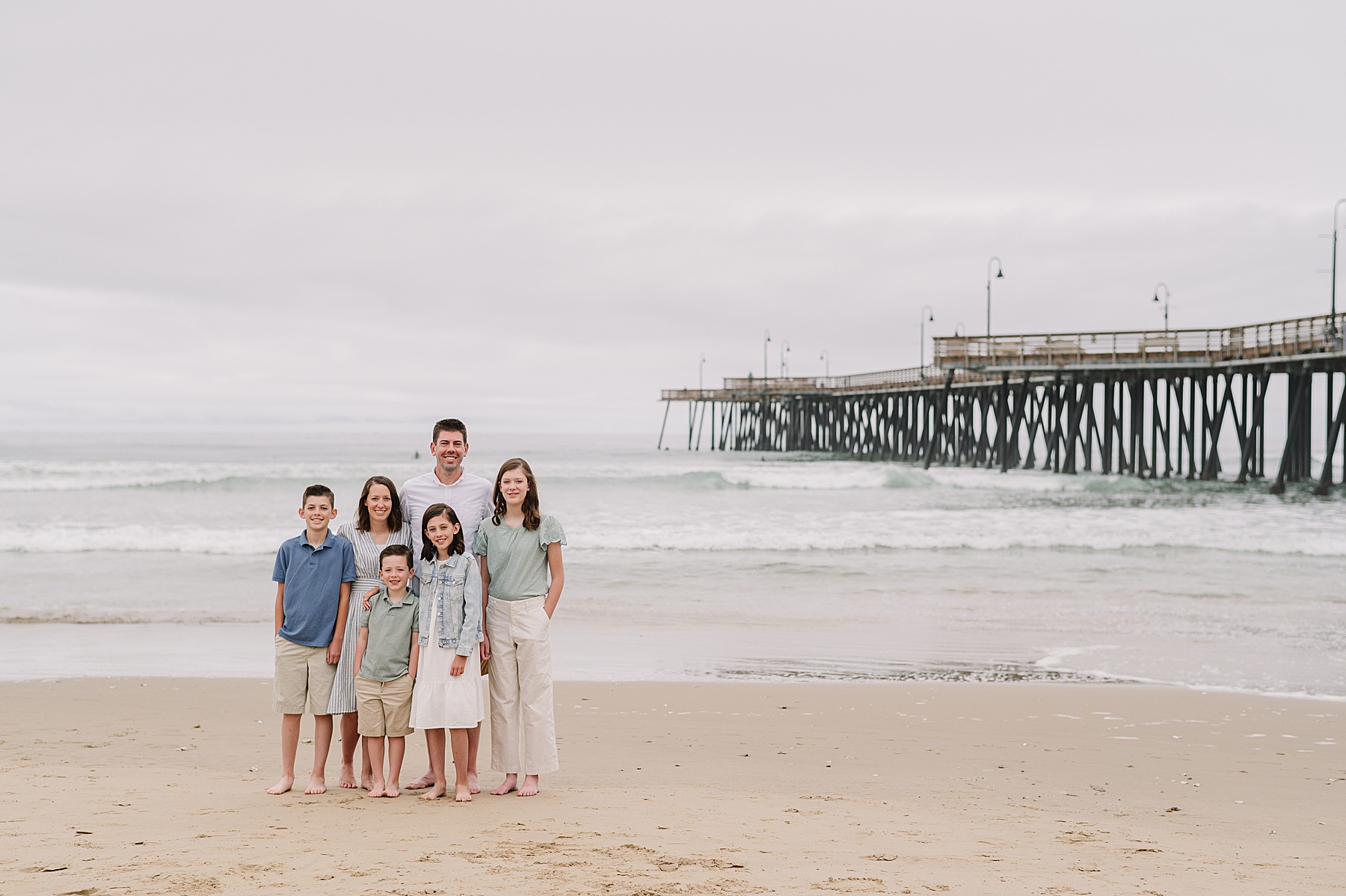 Smiling family posing on the beach with Pismo Beach Pier in the background during their summer family photos in San Luis Obispo.