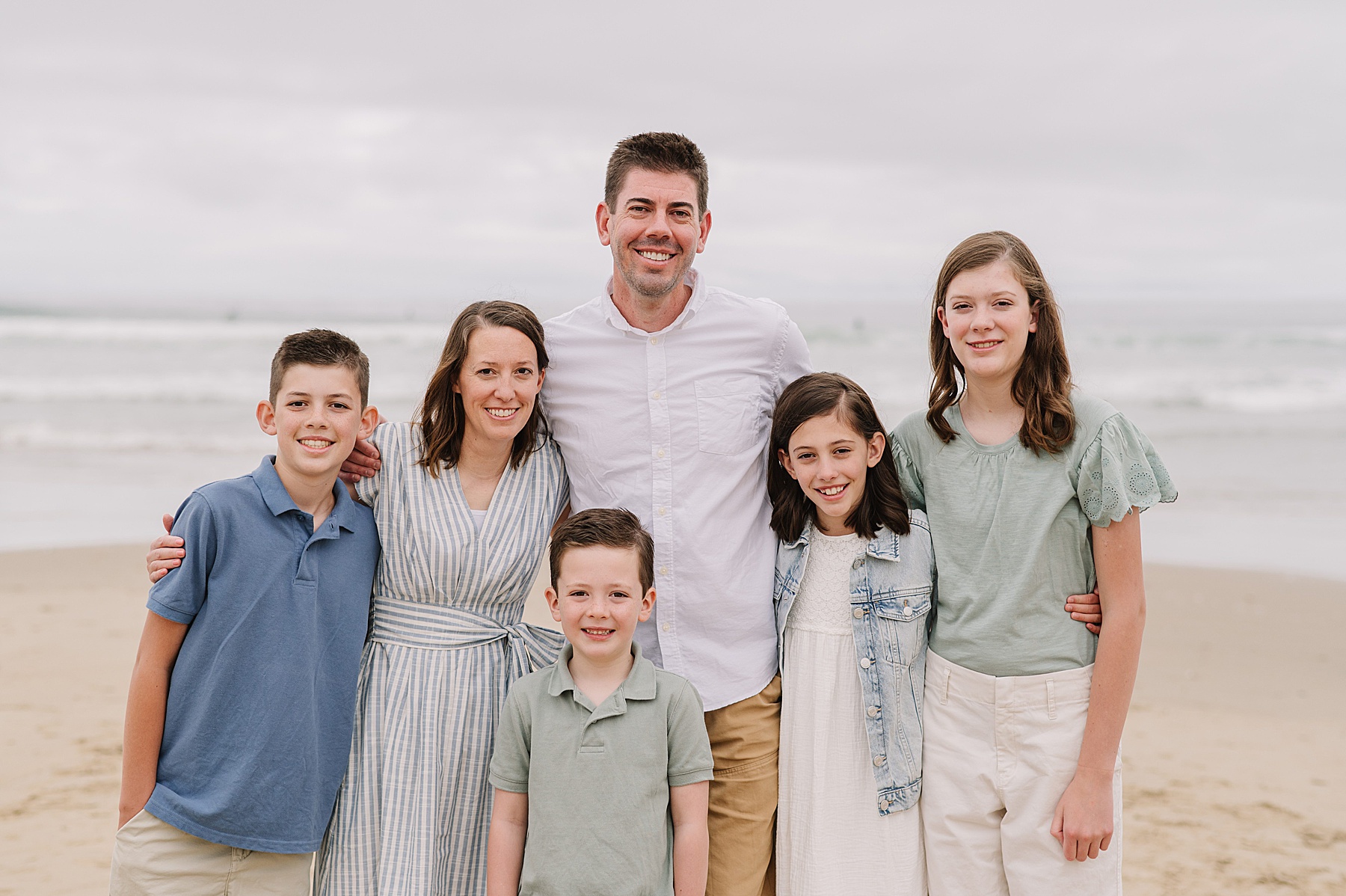 Smiling family of six posing together on the beach during their summer family photos in San Luis Obispo, with soft waves and overcast skies in the background.