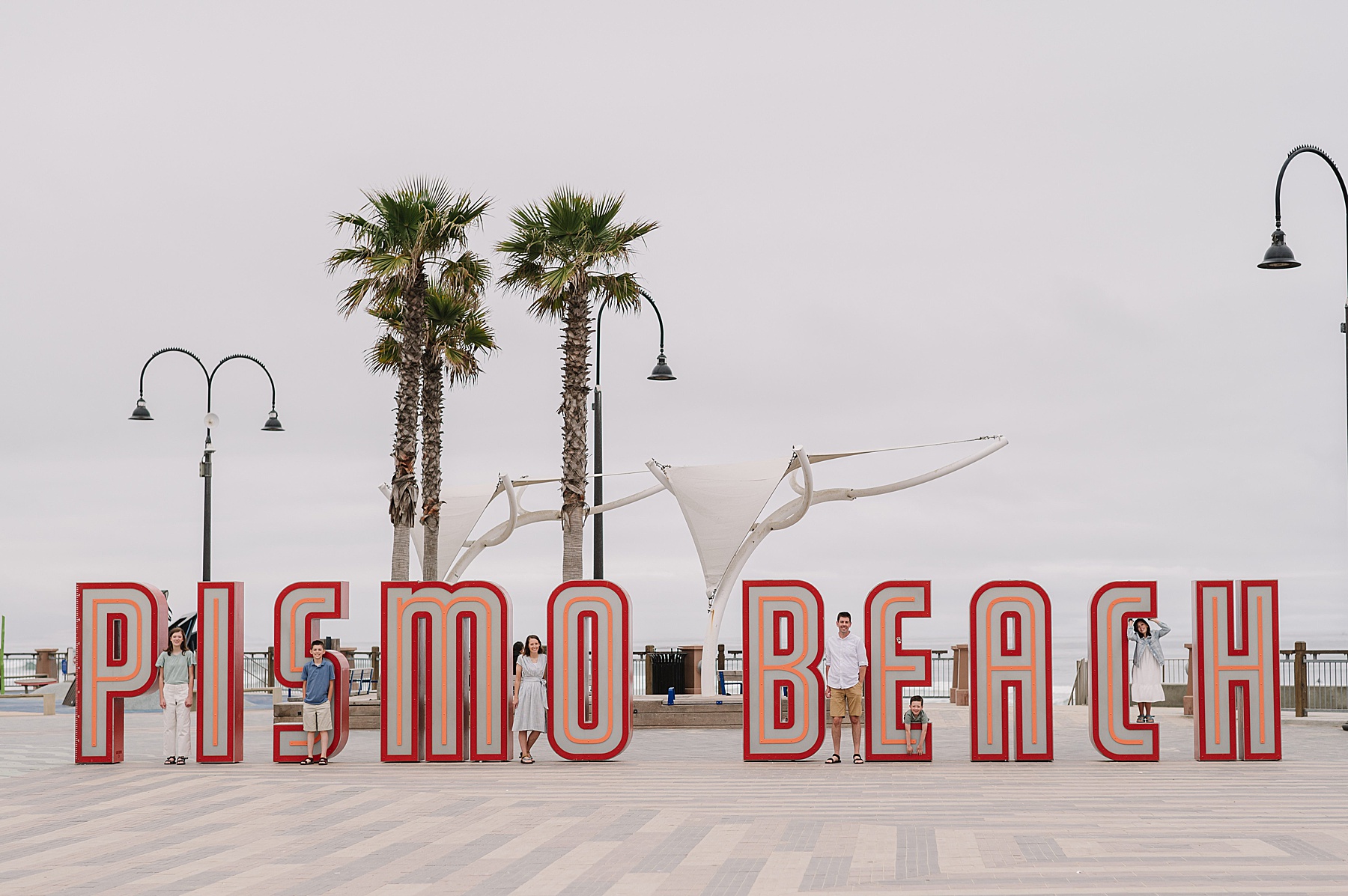 Family photos in San Luis Obispo featuring a playful group posing with the large “Pismo Beach” sign on a cloudy day near the oceanfront boardwalk.