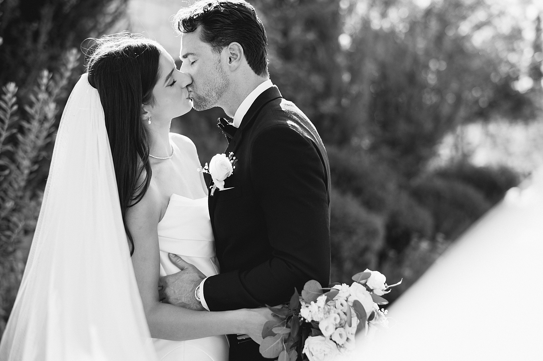 A black and white photo of a bride and groom sharing a romantic kiss on their wedding day. The bride wears a strapless gown with a long veil, and the groom is in a classic tuxedo with a white boutonniere. She holds a bouquet of fresh white flowers.