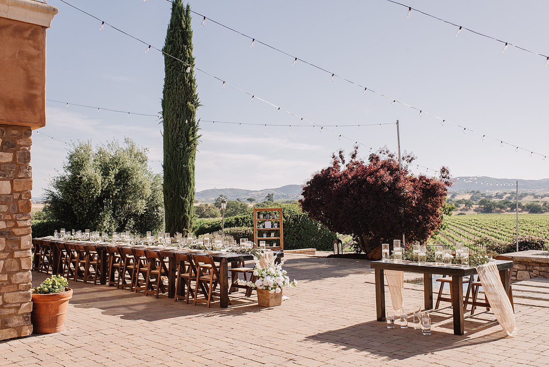 An elegant outdoor reception setup at Mira Vista Estate featuring long wooden tables adorned with candles and greenery, strung market lights overhead, and sweeping vineyard views in the background.