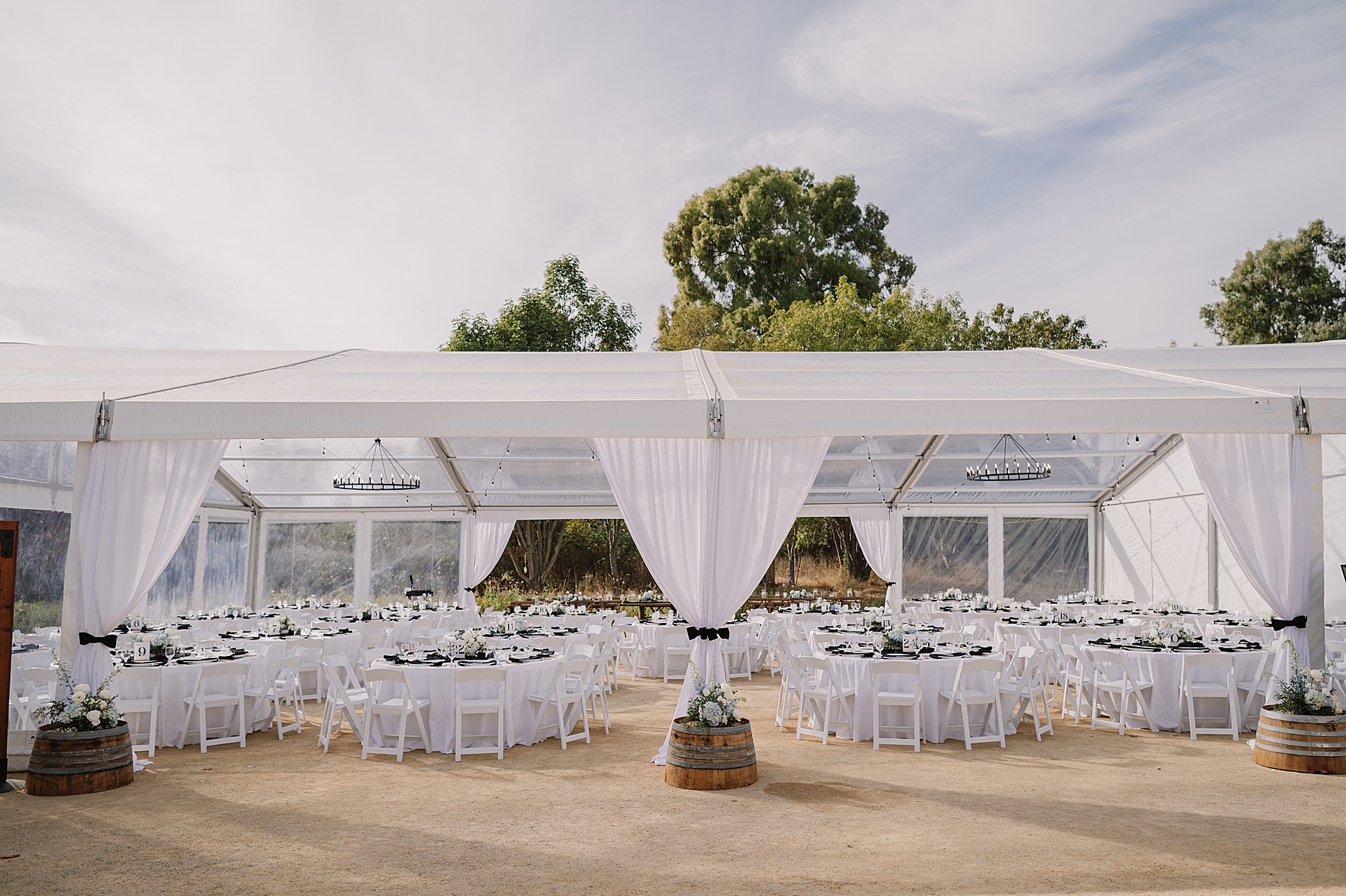 Wedding reception setup at Tar Creek Ranch under a clear-top tent, featuring round tables with white linens, black napkins, and floral centerpieces, framed by flowing white drapery and wine barrel decor.