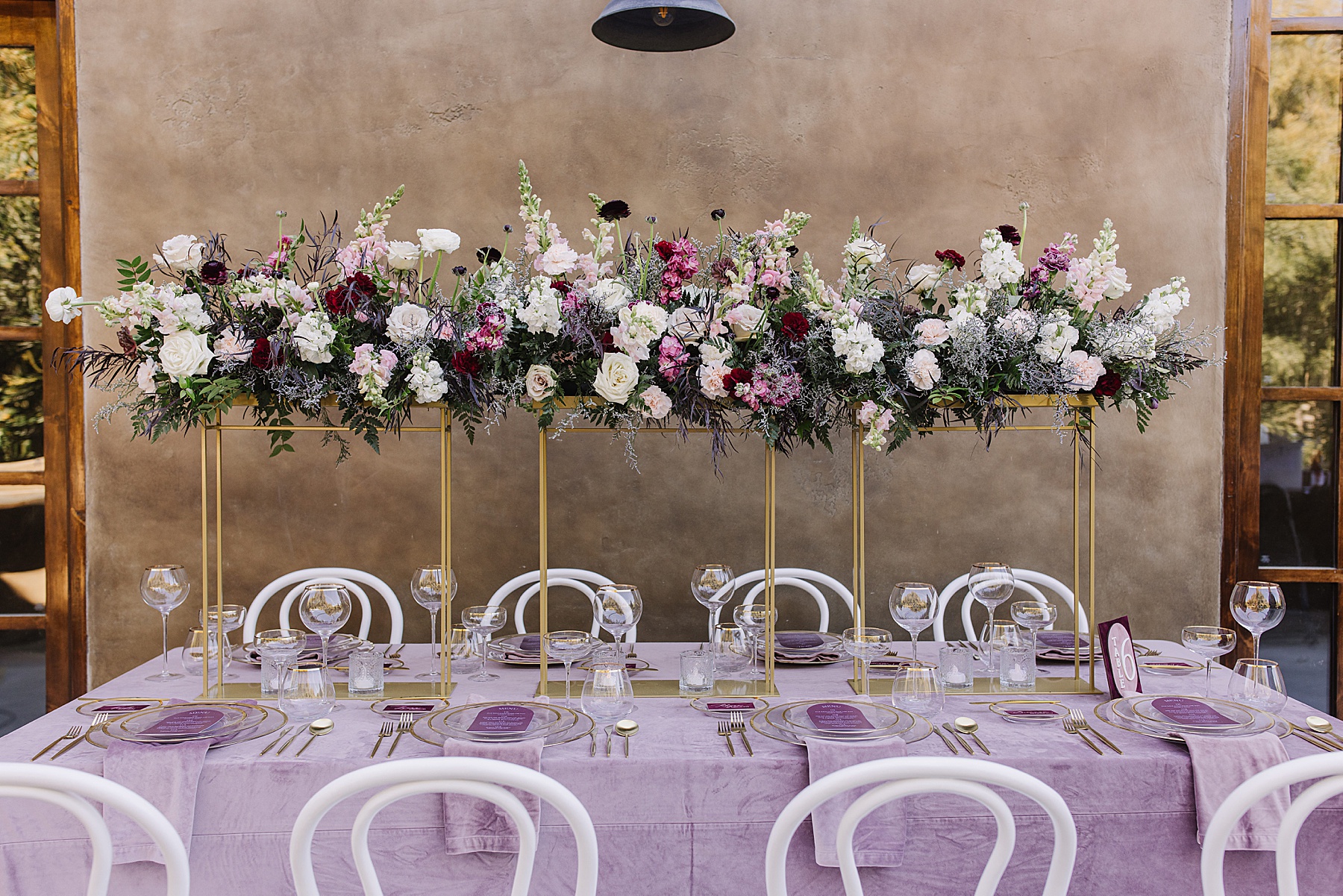 Elegant wedding reception table setup at Villa Loriana, featuring lavender velvet linens, white chairs, gold flatware, and tall gold floral stands with lush pink, burgundy, and white flower arrangements.