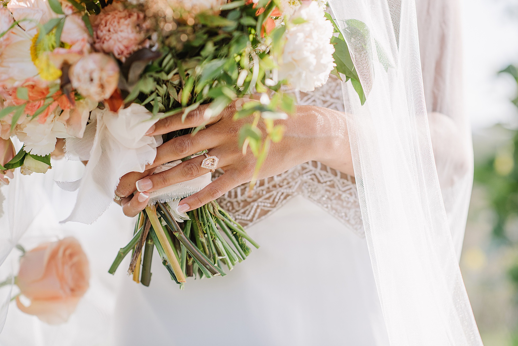 Close-up of a bride’s hand holding a vibrant bridal bouquet at La Lomita Ranch, featuring soft pink and peach florals, trailing greenery, and a delicate white ribbon. The bride wears a diamond engagement ring and a dress with beaded detailing beneath a sheer veil.