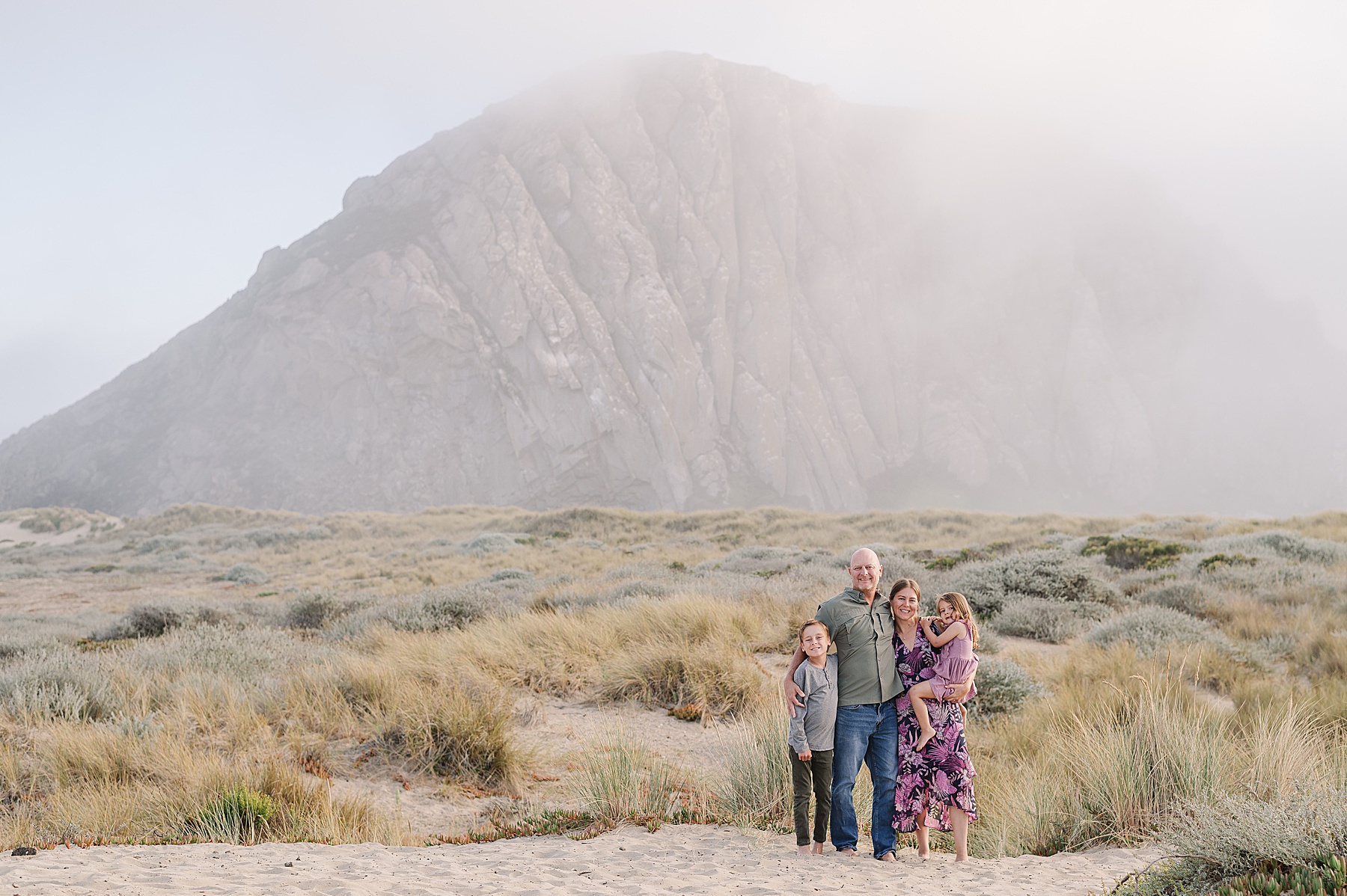 A family of four stands smiling on a sandy beach in front of a large, fog-covered rock formation at Morro Bay. The parents stand in the center with the mother holding her daughter on one side and their son standing on the other. Coastal grasses and misty light add a soft, natural atmosphere