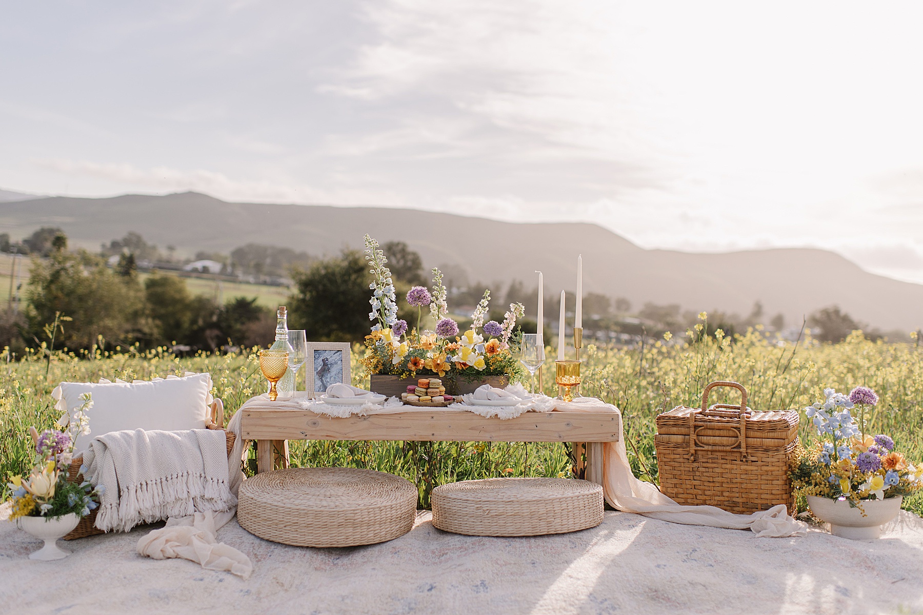 “A styled picnic setup in a wildflower field at golden hour featuring low wooden table décor, woven cushions, candles, florals, and a picnic basket, offering inspiration for creative proposal ideas.”