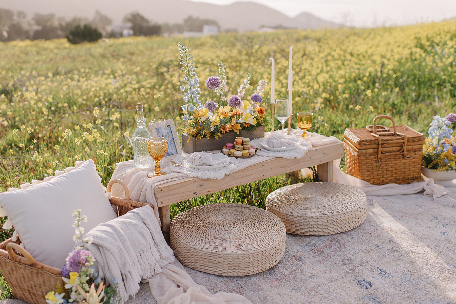 A romantic picnic setup in a wildflower field featuring a rustic low wooden table adorned with vibrant spring florals, gold-rimmed glasses, elegant candles, and French macarons—an intimate scene perfect for unique proposal ideas.
