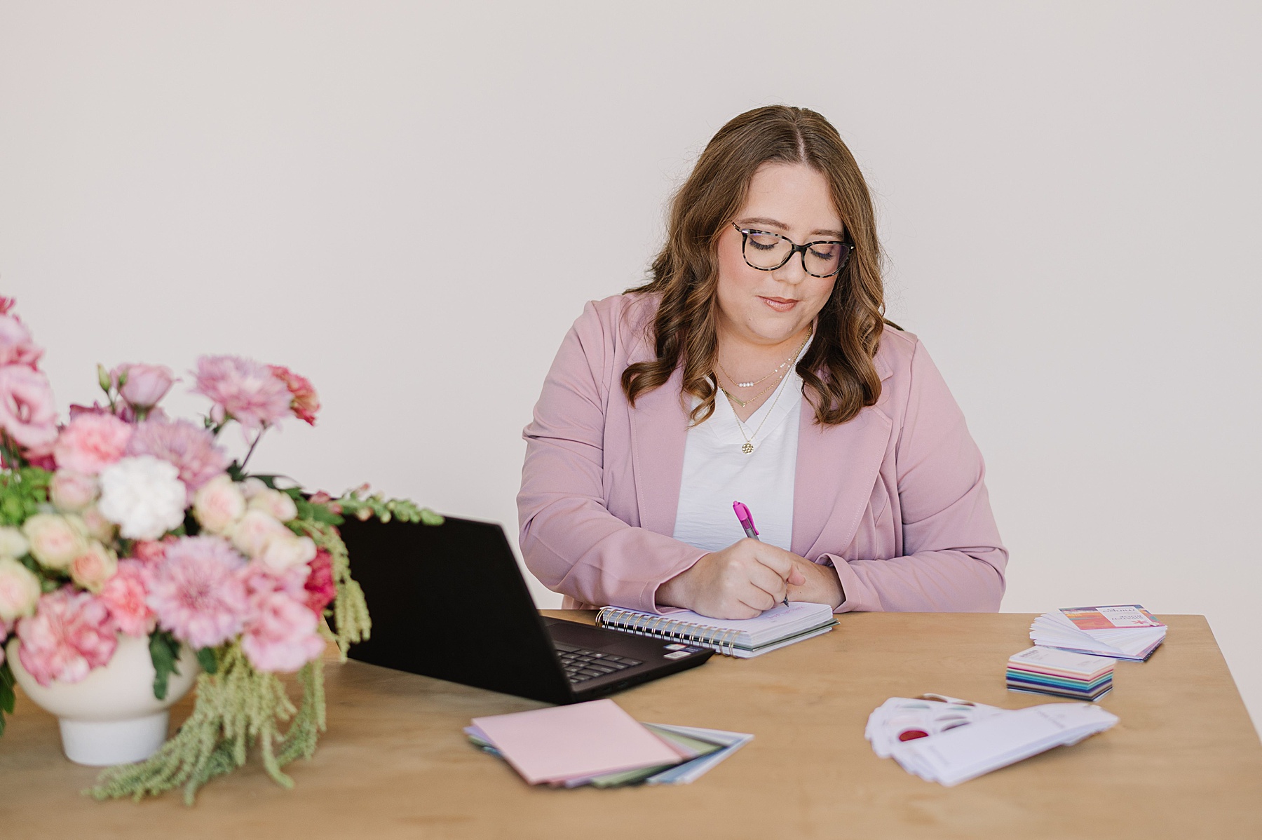 Woman in a pink blazer writing in a notebook during a branding photoshoot. She is seated at a wooden desk with a laptop, colorful swatches, and a vibrant pink floral arrangement, creating a bright and professional workspace scene.
