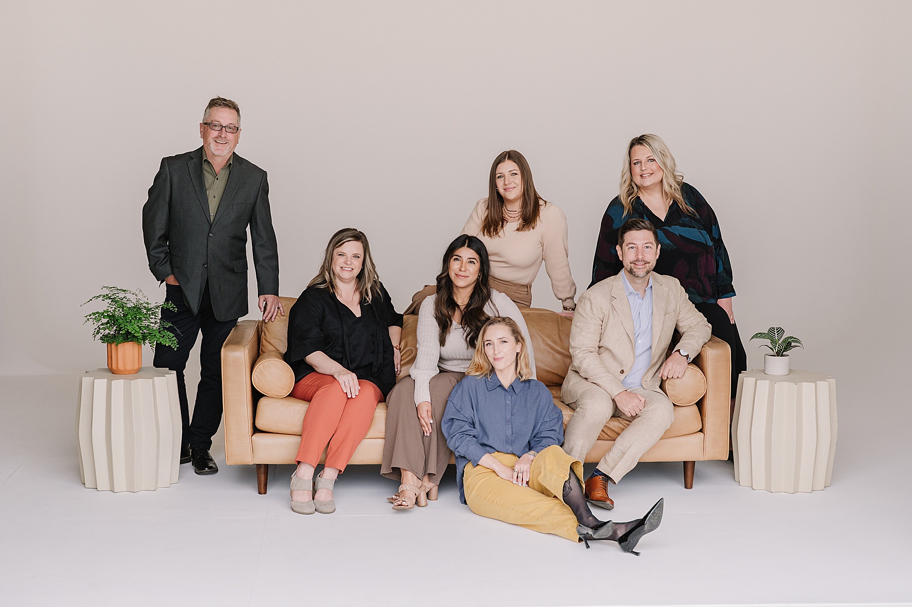 A professional team branding photo taken in a minimalist studio. The group of seven adults—four women seated on a camel-colored leather couch, two women and one man standing behind them, and one man sitting on the armrest—are dressed in stylish business casual attire. Flanking the couch are two modern cream-colored side tables with potted plants. The vibe is friendly, welcoming, and polished.