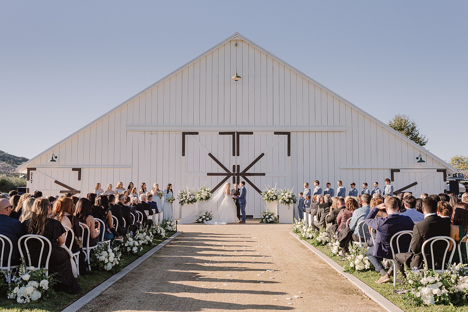 Outdoor ceremony at White Barn Wedding Venue in Edna Valley, California featuring a large white barn, elegant white floral arrangements, and guests gathered for a romantic Central Coast wedding.