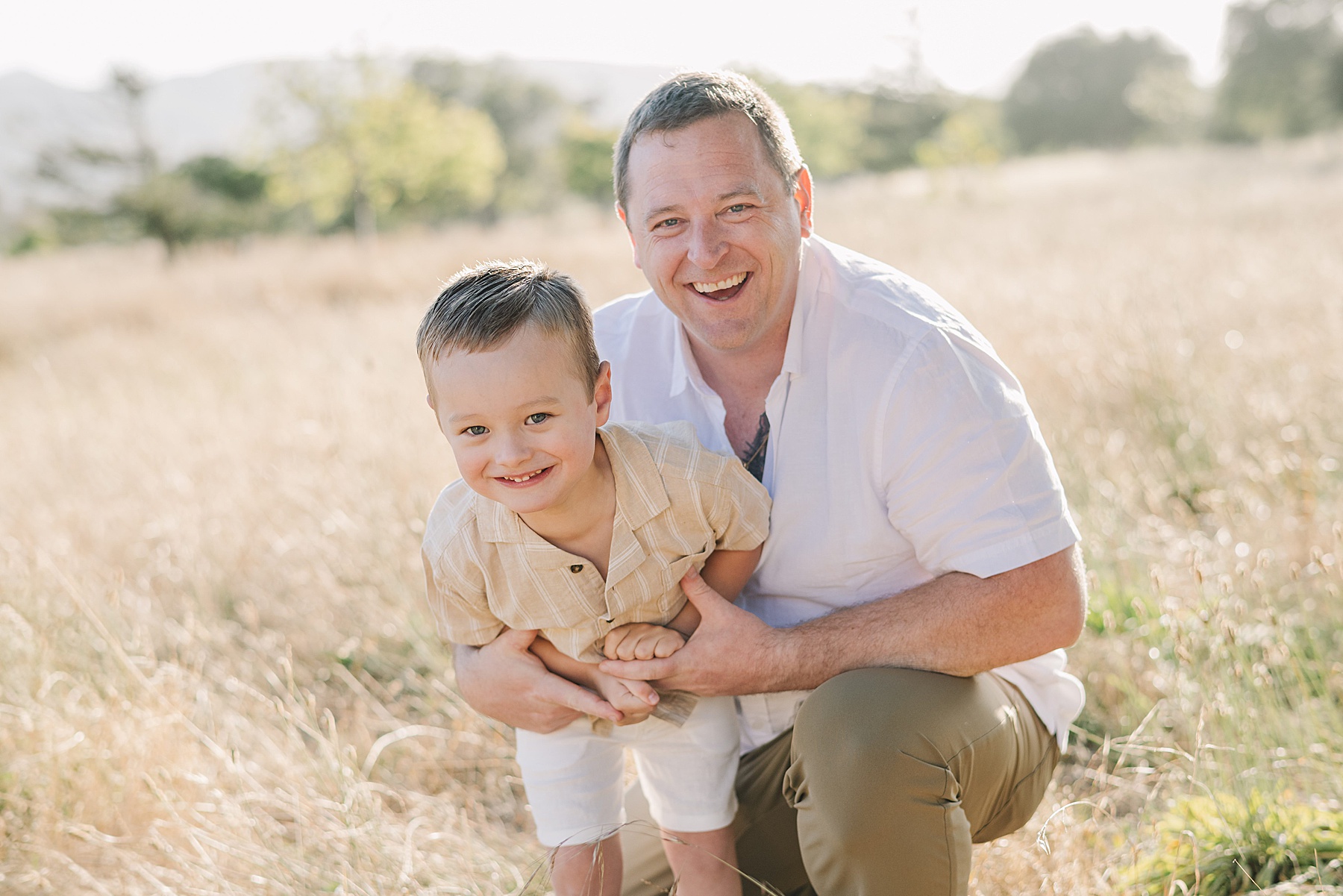 Family photos of a father and young son smiling together in a golden field during an outdoor photography session. The father kneels beside his son as they laugh together, creating a warm, candid family portrait in natural light.