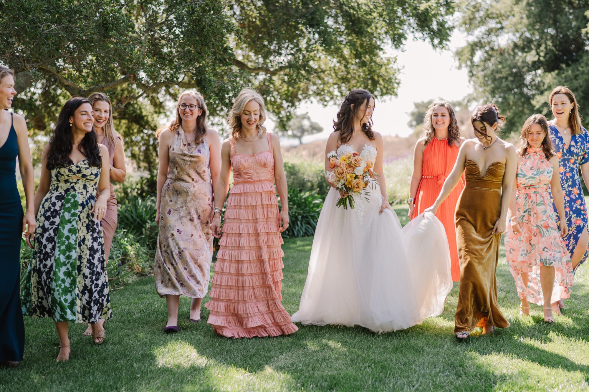 Group of women in elegant, colorful dresses walking outdoors on a sunny day, with the bride in a white gown holding a vibrant bouquet of flowers; a beautiful moment that captures the essence of 2025 wedding trends focused on bold colors, natural backdrops, and personalized styles.