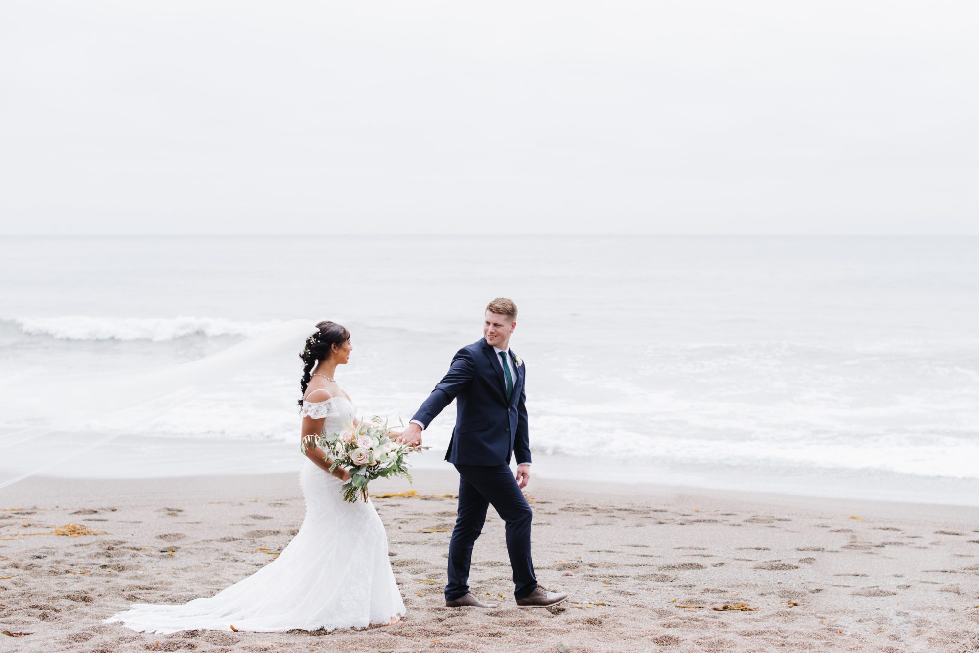 Beautiful California beach wedding photo. Bride and groom are walking next to the ocean. Bride is wearing a wedding dress and holding a bouquet.
