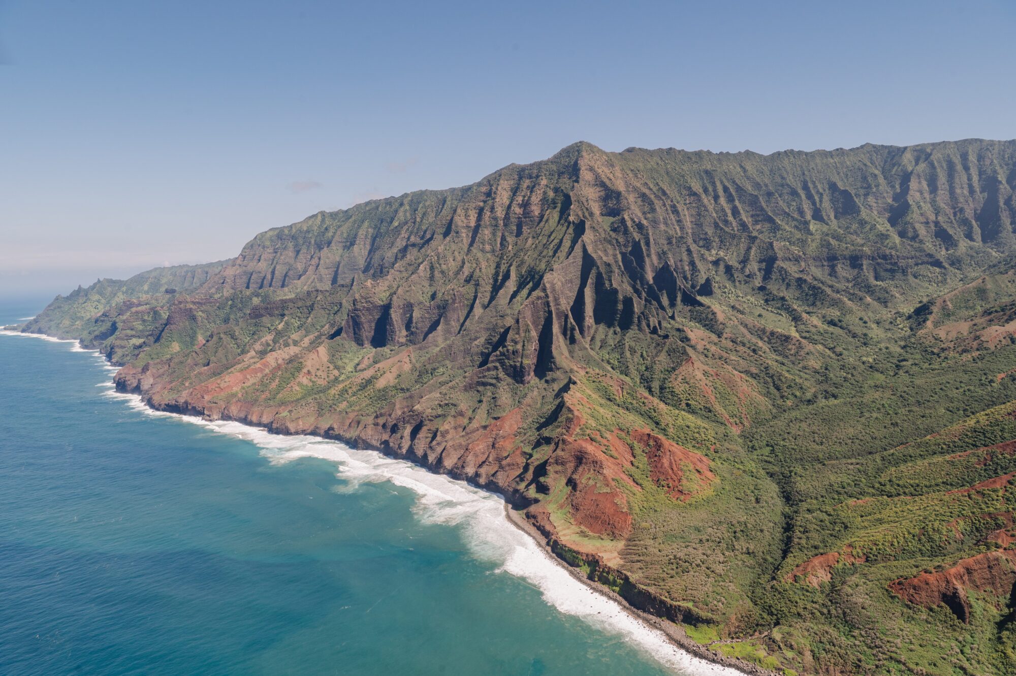 Stunning aerial view of the rugged Napali Coast on Kauai, Hawaii, showcasing dramatic green cliffs, deep valleys, and the blue Pacific Ocean meeting the shoreline.